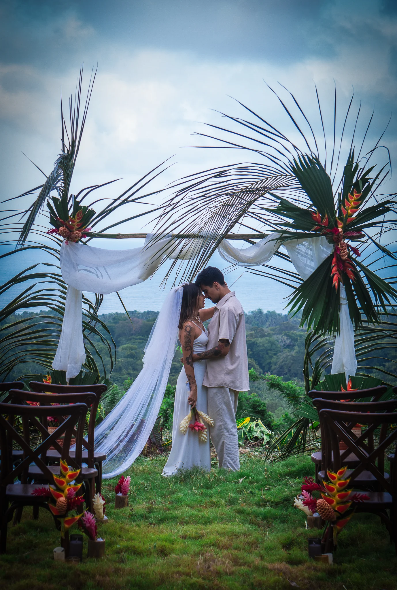 Pareja de novios durante una ceremonia de boda al aire libre en Tayrona, bajo un arco tropical decorado con hojas de palma y flores exóticas.